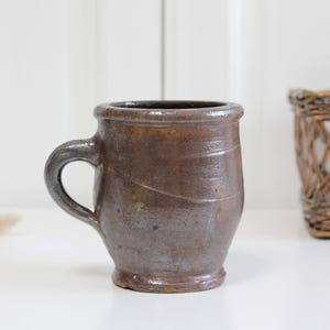 Puede incluir: Una taza de cerámica de color marrón oscuro con un acabado de esmalte brillante y moteado. La taza presenta un borde grueso y redondeado, un asa curva resistente y un cuerpo ligeramente cónico con sutiles estrías horizontales.