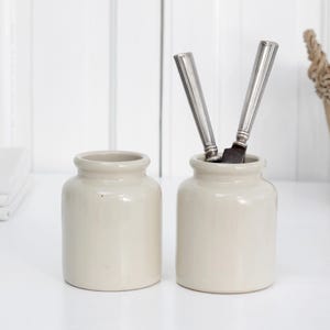 May include: Two cream-coloured ceramic jars, one holding two silver knives. A stack of white folded linens and a wicker basket are in the background. The jars are on a white surface.