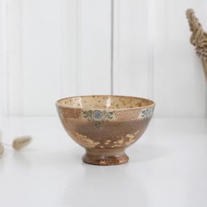 Puede incluir: Un cuenco de cerámica con pie y esmalte moteado en tonos beige y marrón. El borde exterior presenta una banda decorativa con patrones geométricos repetitivos en color marrón rojizo y pequeños motivos florales azules.