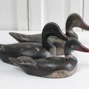 May include: Three hand-carved wooden duck decoys featuring dark black bodies with weathered gray and white underbellies. Each bird has a painted red beak, a defined neck ring, and visible carved feather details along the wings and back.