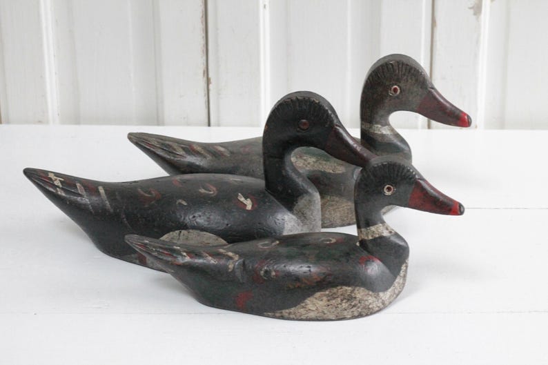 May include: Three hand-carved wooden duck decoys featuring dark black bodies with weathered gray and white underbellies. Each bird has a painted red beak, a defined neck ring, and visible carved feather details along the wings and back.