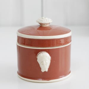 Puede incluir: Un tarro cilíndrico de cerámica color terracota con tapa. El tarro presenta un borde blanco y un relieve blanco en relieve de una cara en la parte delantera. La tapa tiene un pomo blanco. Un recipiente de almacenamiento decorativo.