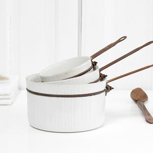 Puede incluir: Un conjunto de cacerolas de esmalte blanco con asas y bordes de metal marrón. Una cuchara de madera descansa cerca. Las cacerolas están anidadas, de diferentes tamaños. El fondo es una pared de madera blanca.