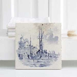 May include: A square ceramic tile featuring a blue and white monochromatic illustration of a harbour scene with sailing ships, a windmill, and buildings along the shoreline. The detailed maritime artwork is centred on the off-white tile surface.