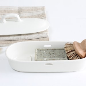 Könnte beinhalten: Weiße Keramik-Schale mit einem Stück Seife und einer Holzbürste. Die Schale hat einen Deckel mit einem Griff. Die Seife ist grün und trägt den Text "Savon de Marseille" und "Provence".