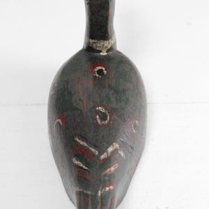 May include: A hand-carved wooden duck decoy featuring a dark brown base coat with weathered green, red, and white painted accents. The bird figure displays a slender neck, a rounded body, and stylized feather patterns painted along the back and tail.