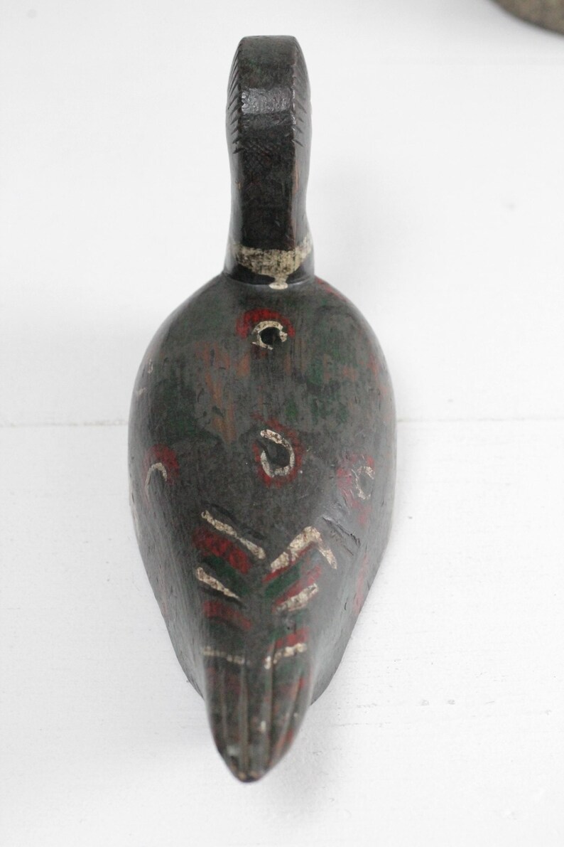 May include: A hand-carved wooden duck decoy featuring a dark brown base coat with weathered green, red, and white painted accents. The bird figure displays a slender neck, a rounded body, and stylized feather patterns painted along the back and tail.