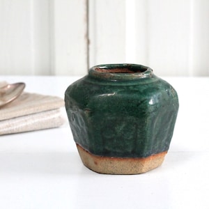 May include: A small, antique green ceramic vase with a textured surface and a light brown base. The vase has a rounded shape and a narrow opening. A silver spoon rests on a folded beige cloth.