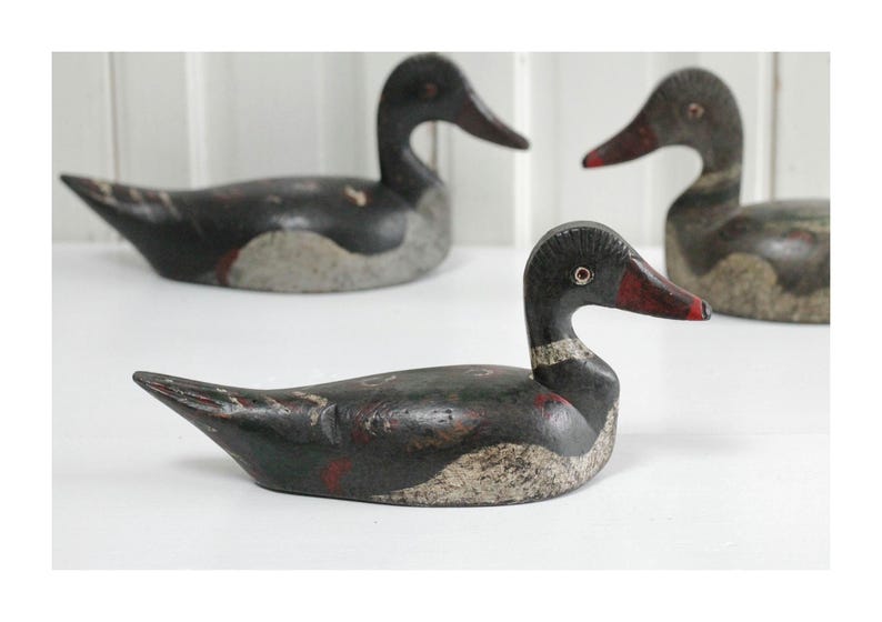 May include: Three hand-carved wooden duck decoys featuring weathered black, gray, and red paint. Each bird has a distinctively shaped head, a pointed tail, and a textured finish showing signs of age and wear across the wooden surfaces.