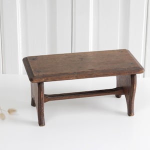 May include: A small rectangular wooden stool with a dark brown finish, featuring a flat top surface and four sturdy legs connected by a horizontal support bar. The wood grain is visible across the surface, showing natural texture and wear.