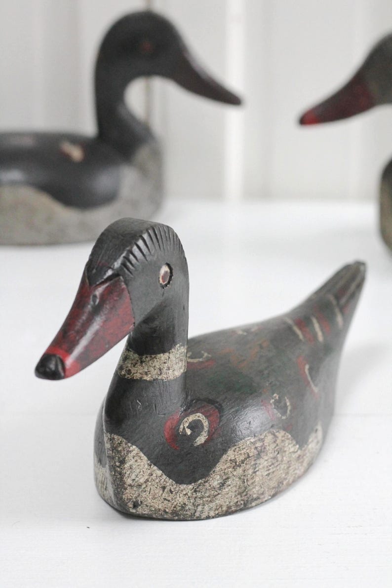 May include: A hand-carved wooden duck decoy featuring a dark black body with distressed white painted accents on the lower chest and wings. The duck has a red beak with a black tip, small circular eyes, and decorative red and white circular markings.