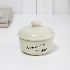 Puede incluir: Tarro de cerámica color crema con tapa y un pequeño pomo en la parte superior. El tarro tiene las palabras "Spécialité MAISON" en escritura negra. El tarro está sobre una superficie blanca.