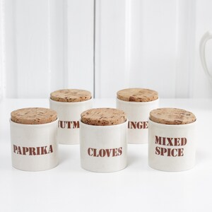 Op de afbeelding: Vijf witte keramische kruidenpotten met kurken deksels, met de labels "Paprika", "Kruidnagel", "Gemengde specerijen" en twee andere. Een witte kan en een houten lepel liggen op een wit oppervlak.