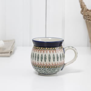Może przedstawiać: Ceramiczny kubek z niebieską obwódką i uchwytem, ​​z wzorem zielonych, czerwonych i złotych elementów na białym tle. Kubek stoi na białej powierzchni, z koszem i innymi przedmiotami w tle.