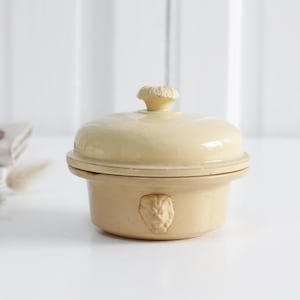 Könnte beinhalten: Eine kleine, cremefarbene Keramik-Auflaufform mit Deckel und Löwenkopf-Detail. Die Schüssel ist rund mit einem kleinen Griff oben. Die Schüssel steht auf einer weißen Oberfläche.