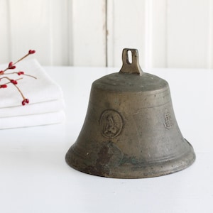 May include: Antique bronze bell with a flared base and a loop handle. The bell has embossed designs and a weathered patina. A sprig of red berries rests on a stack of white folded linens.