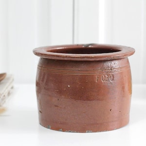 May include: A brown cylindrical stoneware crock with a wide rim and a flat base. The exterior surface features a smooth, glazed finish with the embossed text F 0.70 visible on the side. The vessel has a thick, rounded top edge and a sturdy construction.