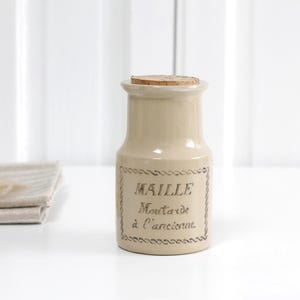 Puede incluir: Un tarro de mostaza de cerámica beige con tapón de corcho. El tarro tiene una etiqueta rectangular con el texto "MAILLE Moutarde à l'ancienne" en una fuente decorativa. A la izquierda del tarro hay una servilleta de lino beige doblada.