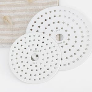 Pode incluir: Duas bases para panelas circulares de cerâmica branca com numerosos orifícios pequenos. As bases para panelas são de tamanhos diferentes e têm um orifício central. A base para panelas maior tem o texto "4263" e "WIZA-BPL". Um tecido listrado bege e branco no fundo.