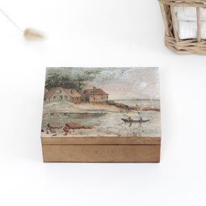 May include: A rectangular wooden box featuring a painted landscape on its lid. The artwork depicts two houses situated on a grassy shoreline near a body of water, with a small boat carrying two figures floating on the surface under a soft sky.