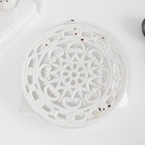 May include: White cast iron trivet with an ornate, openwork design. A black ladle rests beside the trivet. Two white ceramic salt and pepper shakers and a stack of white napkins are also in the image.