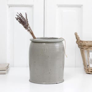 Puede incluir: Un jarrón de gres gris con un ramo de lavanda seca. El jarrón cilíndrico rústico tiene un borde redondeado y una cuerda clara. Una cesta de mimbre y ropa de cama doblada también están en la imagen.