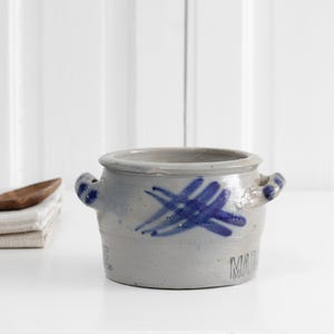 Peut inclure: Un pot en céramique gris clair doté de deux anses latérales et d'un motif en croix peint à la main en bleu sur le devant. La base du récipient présente des lettres en relief partielles. Le pot a une large ouverture et une finition émaillée.