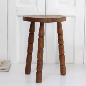 May include: A three-legged wooden stool with a round seat and turned legs. The stool features a dark brown wood grain finish and a simple construction with cylindrical, segmented legs that taper slightly toward the floor.