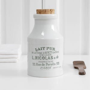 Puede incluir: Una botella de leche de cerámica blanca con tapón de corcho. La botella tiene texto gris que dice "LAIT PUR" y "L. NICOLAS". Una pequeña espátula de madera está al lado de la botella. Una pila de paños blancos doblados está a la izquierda.