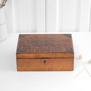May include: A rectangular wooden box with a rich brown grain pattern, featuring dark metal decorative corner accents and a small central keyhole on the front panel. The box has a smooth, flat lid and a solid construction, presenting a classic appearance.