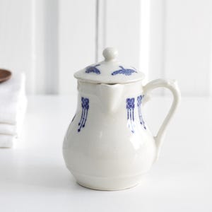 May include: A white ceramic creamer with a lid, decorated with blue floral patterns. The creamer has a curved handle and a spout. A stack of white towels and a wooden spoon are in the background.