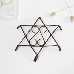 Może przedstawiać: Brązowy metalowy podkładka pod garnek w kształcie gwiazdy Dawida. Podkładka ma geometryczny wzór z zakrzywionymi akcentami. Zaprojektowana do ochrony powierzchni przed gorącymi naczyniami.