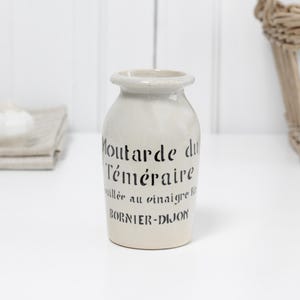 以下が含まれることがあります： 「Moutarde du Téméraire」と「Bornier-Dijon」と書かれた黒い文字が入ったクリーム色の陶器の瓶。瓶は丸みを帯びたボディと小さなフレアリムを持っています。白い表面に置かれ、背景にはバスケットと装飾的なニンニクの球根があります。