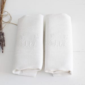 Könnte beinhalten: Zwei weiße Leinen-Handtücher mit gestickten Blumendesigns. Die Handtücher sind gefaltet und auf einer weißen Oberfläche platziert.