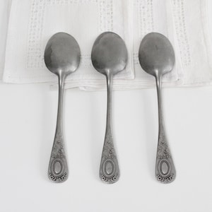 以下が含まれることがあります： 白いテクスチャード加工の布の上に置かれた、装飾的なハンドルが付いた3つのアンティークシルバーのスプーン。スプーンはハンドルの付け根に楕円形のデザインがあります。スプーンは白い背景に対して一列に並んでいます。