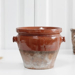 May include: A glazed, antique-style ceramic pot with a rich brown hue. The pot features two small handles and a wide opening. The lower portion has a faded, aged appearance, suggesting it is vintage. A decorative piece for the home.
