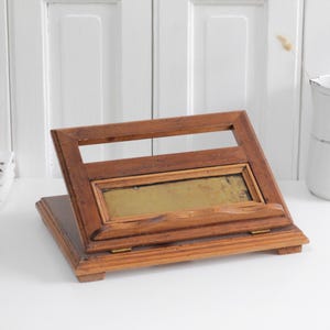 May include: A wooden book stand featuring a rectangular frame with a central brass plate and a hinged base. The stand is constructed from medium-toned wood with visible grain and includes a small ledge at the bottom to support a book or tablet.