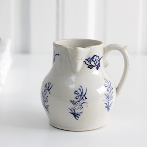 May include: A white ceramic jug with a handle and spout, decorated with blue floral patterns. The jug is on a white surface. A wooden spoon and folded white towels are in the background.