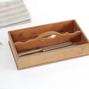 May include: A wooden cutlery tray with a handle, holding a knife and a spoon. A folded beige and white striped linen napkin is in the background. The tray is made of light brown wood.