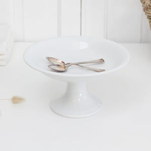 May include: A white ceramic pedestal bowl with a wide, shallow basin and a sturdy base. Two small, metallic silver-toned spoons are resting inside the bowl, positioned in a crossed arrangement. The surface of the bowl is smooth and glossy.