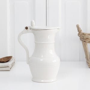May include: A white ceramic jug with a hinged lid, a curved handle, and a wide spout. The jug features a smooth, glossy finish and a rounded body shape, standing upright with a small knob on top of the lid for easy opening and closing.