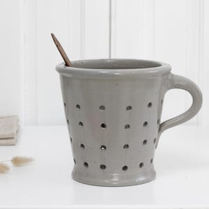 Puede incluir: Una taza de cerámica gris con asa y numerosos agujeros pequeños. Una cuchara de madera descansa dentro de la taza. La taza está sobre una superficie blanca, con un paño doblado y una cesta de mimbre en el fondo.