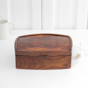 Könnte beinhalten: Eine rechteckige Holzschatulle mit dunkelbrauner Holzmaserung und einem gewölbten Deckel. Die Schatulle verfügt über einen kleinen Metallverschluss in der vorderen Mitte. Das Holz hat eine polierte Oberfläche mit sichtbaren natürlichen Mustern.