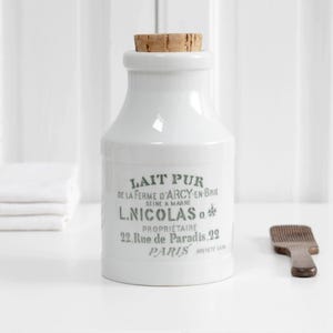 Puede incluir: Un recipiente de cerámica blanca con tapón de corcho, con texto impreso en negro que dice LAIT PUR DE LA FERME D'ARCY EN BRIE SEINE & MARNE L.NICOLAS PROPRIETAIRE 22 Rue de Paradis 22 PARIS BREVETE SGDG, junto a un pequeño peine marrón.