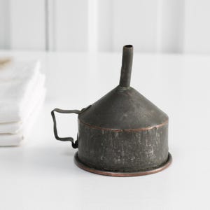 Könnte beinhalten: Ein vintage Metalltrichter mit einem Kupferrand. Der Trichter ist dunkelgrün und hat oben einen Ausguss. Er steht auf einer weißen Oberfläche.