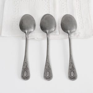 Puede incluir: Tres cucharas de metal plateado dispuestas verticalmente, cada una con un cuenco redondeado y un mango decorado con un patrón floral en relieve y un motivo ovalado central. Las cucharas están colocadas sobre una servilleta de tela blanca.