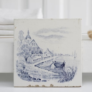 Op de afbeelding: Een vintage blauwe en witte keramische tegel met een schilderachtige dorpscene met huizen, een rivier en een boot. De tegel heeft een rustieke, verweerde uitstraling.