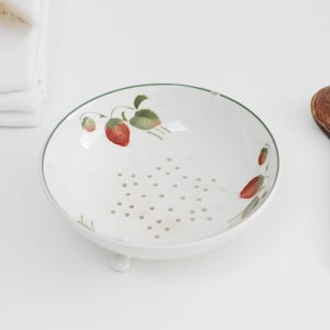 Könnte beinhalten: Eine weiße Keramikschale für Beeren mit einem aufgemalten Erdbeermuster aus grünen Blättern und roten Früchten. Die Schale hat einen perforierten Boden zum Abtropfen, kleine Füße und einen grünen Rand. Daneben liegt ein dunkelbrauner Holzlöffel.