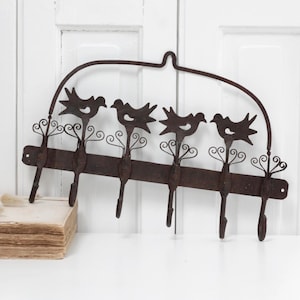 Puede incluir: Un perchero de pared rústico de metal con diseño de pájaros. El gancho marrón tiene cuatro ganchos y adornos decorativos. Los pájaros están silueteados sobre el fondo blanco. El gancho mide unos 46 cm de ancho.