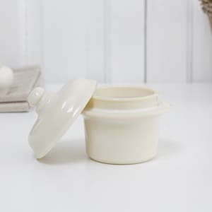May include: Off-white ceramic garlic keeper with a lid, sitting on a white surface. The lid is slightly ajar, revealing the interior. Two ceramic garlic bulbs rest on a linen cloth in the background. A wicker basket is in the background.